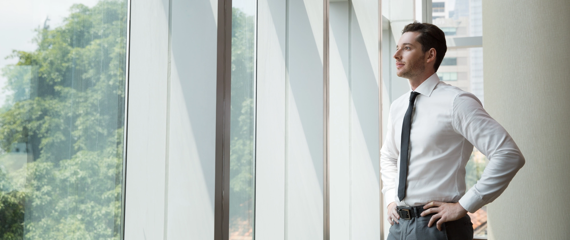 A man in a white dress shirt and tie stands with hands on hips, looking out a large window in a modern office building.