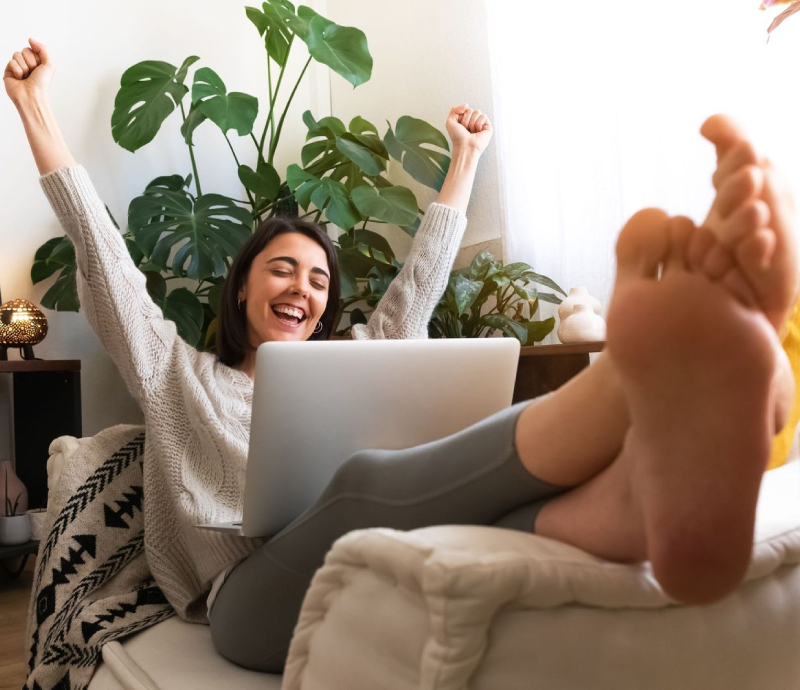 A woman sits on a couch with her feet up, smiling and raising her arms in excitement while using a laptop. There are green plants and natural light in the background.