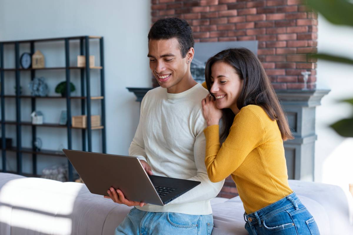 Two people smile while looking at a laptop in a modern living room with shelves and a brick fireplace in the background.