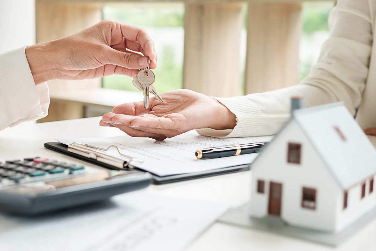 A person hands over keys to another person across a desk with documents, a pen, a calculator, and a small model house.