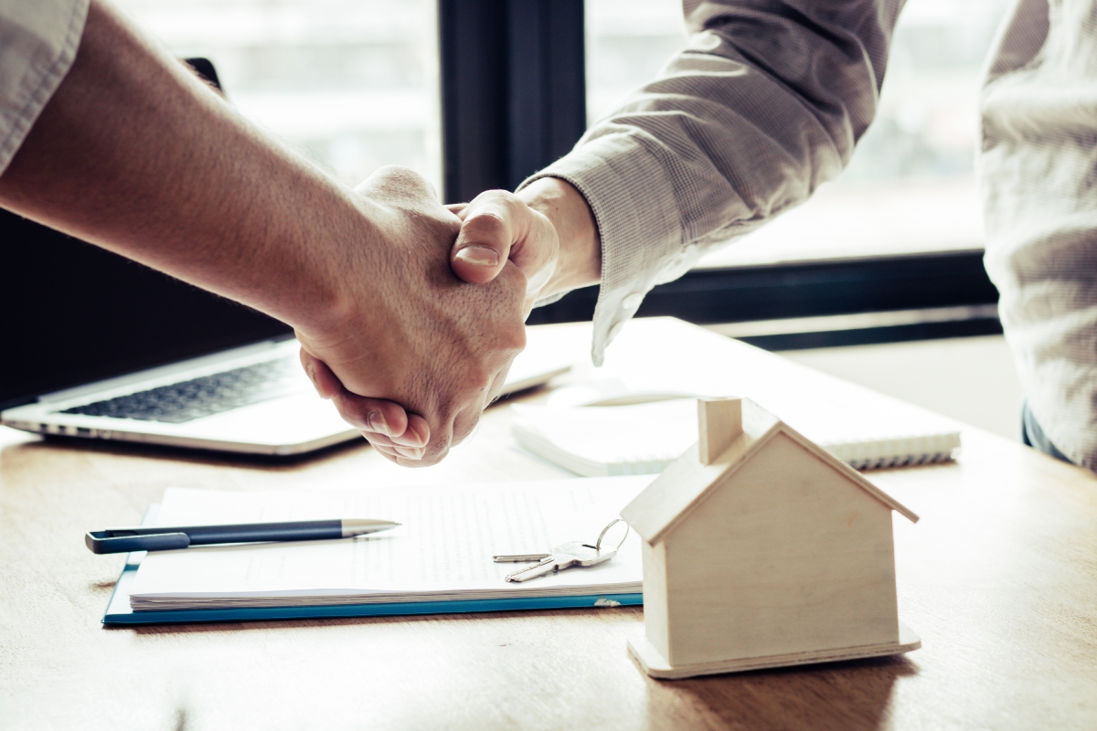 Two people shake hands over a desk with a notebook, pen, house model, and keys, suggesting a real estate or property agreement.