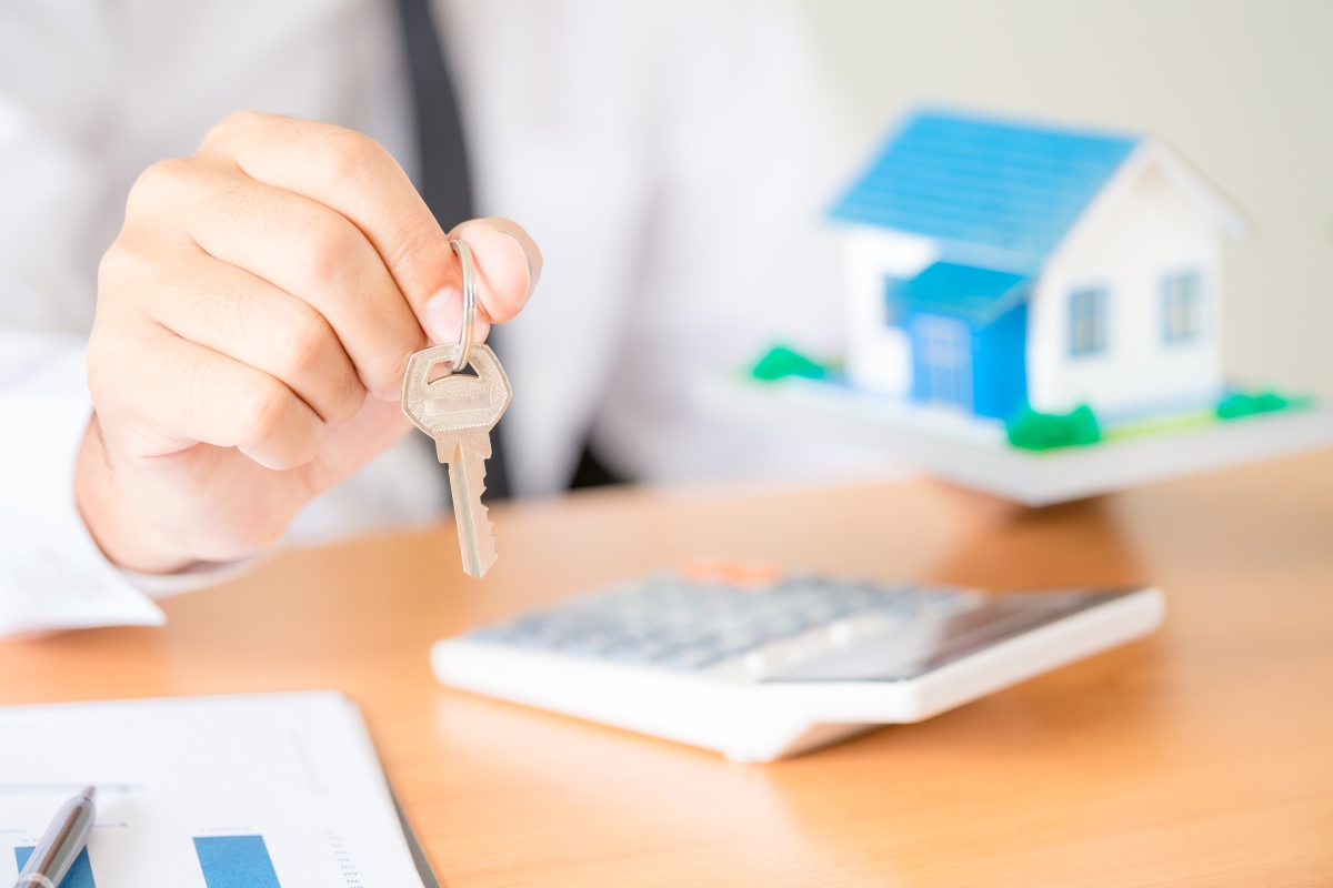 A person holding a key, with a calculator and a small model house on a desk, suggesting a real estate or home buying context.
