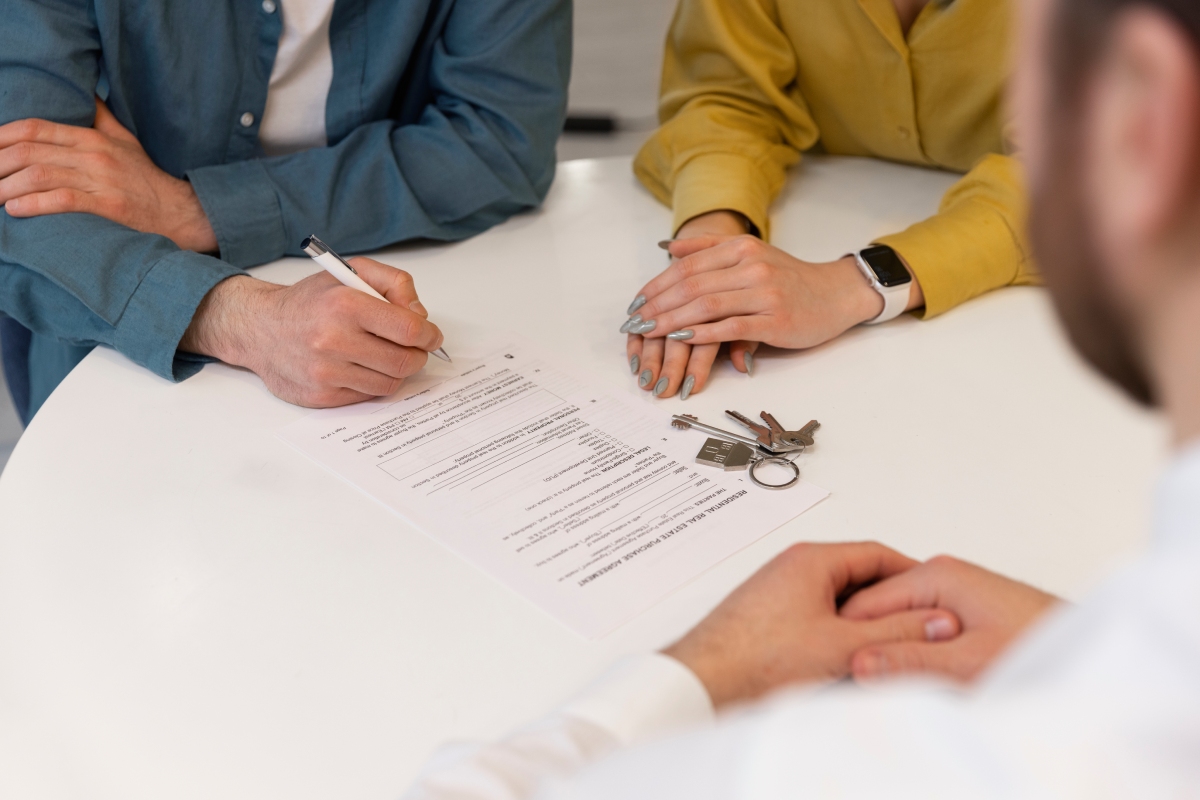 Three people sit at a table with a contract and house keys; one person is signing the document while the others observe.