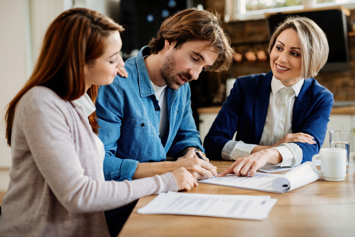 Three people sit at a table reviewing and signing documents, with one woman pointing at the papers and another watching attentively.