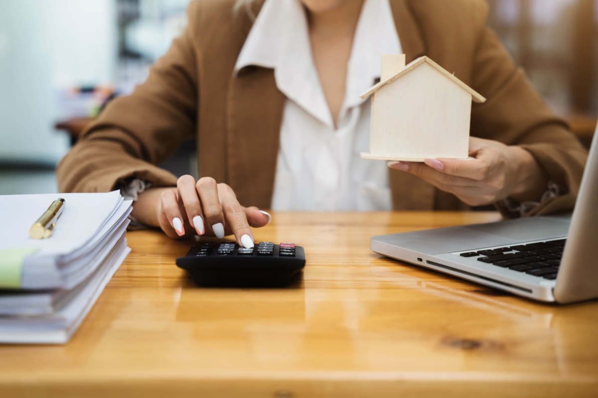 Person in business attire holds a small wooden house model in one hand and uses a calculator with the other, beside a laptop and stack of papers on a wooden desk.
