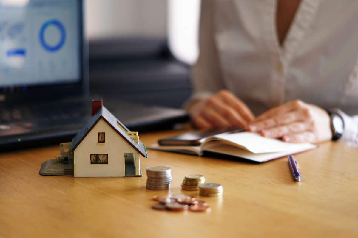 A person sits at a desk with a model house, stacked coins, a notebook, and a laptop displaying a chart.