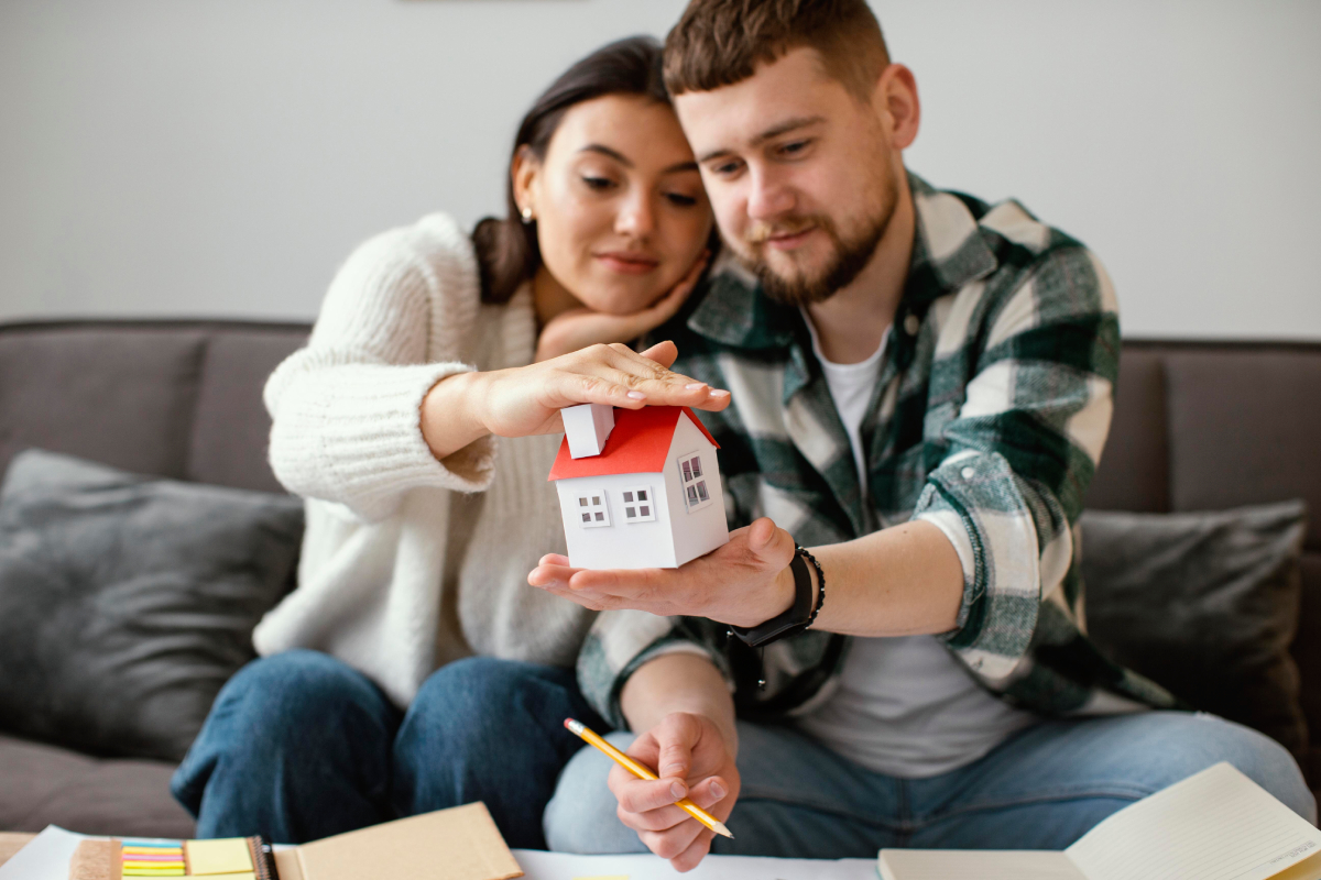 A couple sits on a couch holding a small model house, with papers, a notebook, and a pencil on the table in front of them.