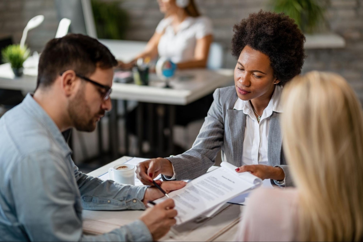 A woman in business attire explains a document to a man and woman seated at a desk, with office workers in the background.