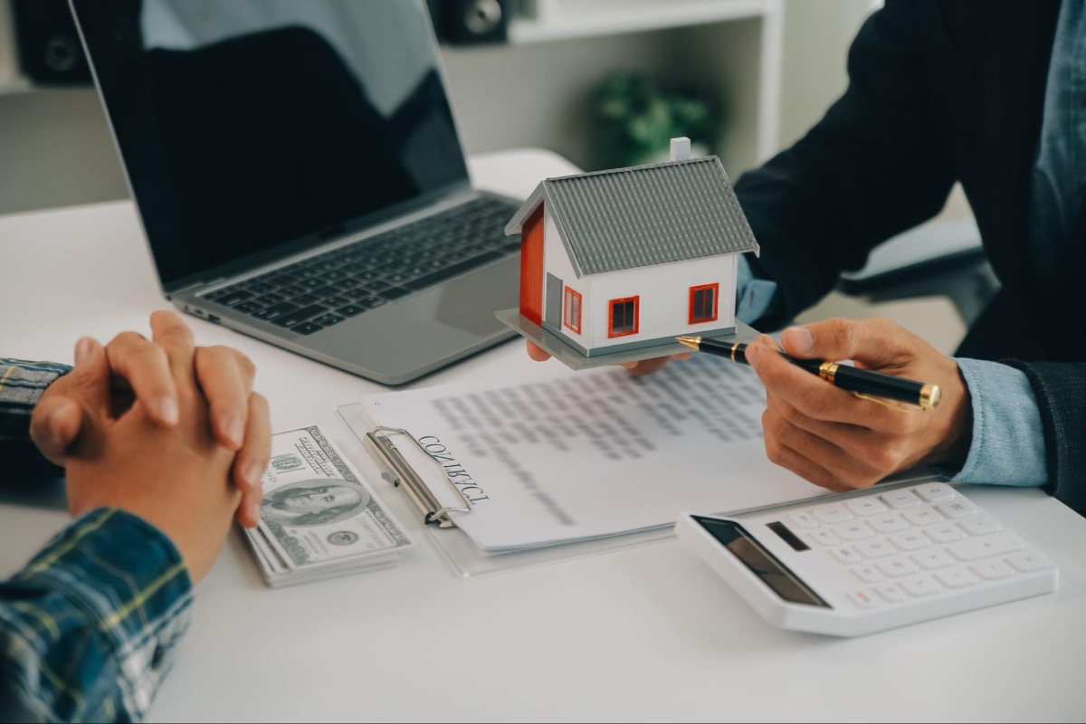 Two people discuss a real estate contract at a desk with a model house, laptop, calculator, stack of cash, and clipboard with contract.