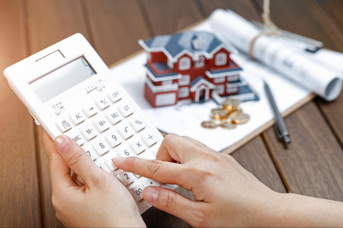 Hands using a calculator next to a clipboard with house model, rolled documents, coins, and a pen on a wooden table.