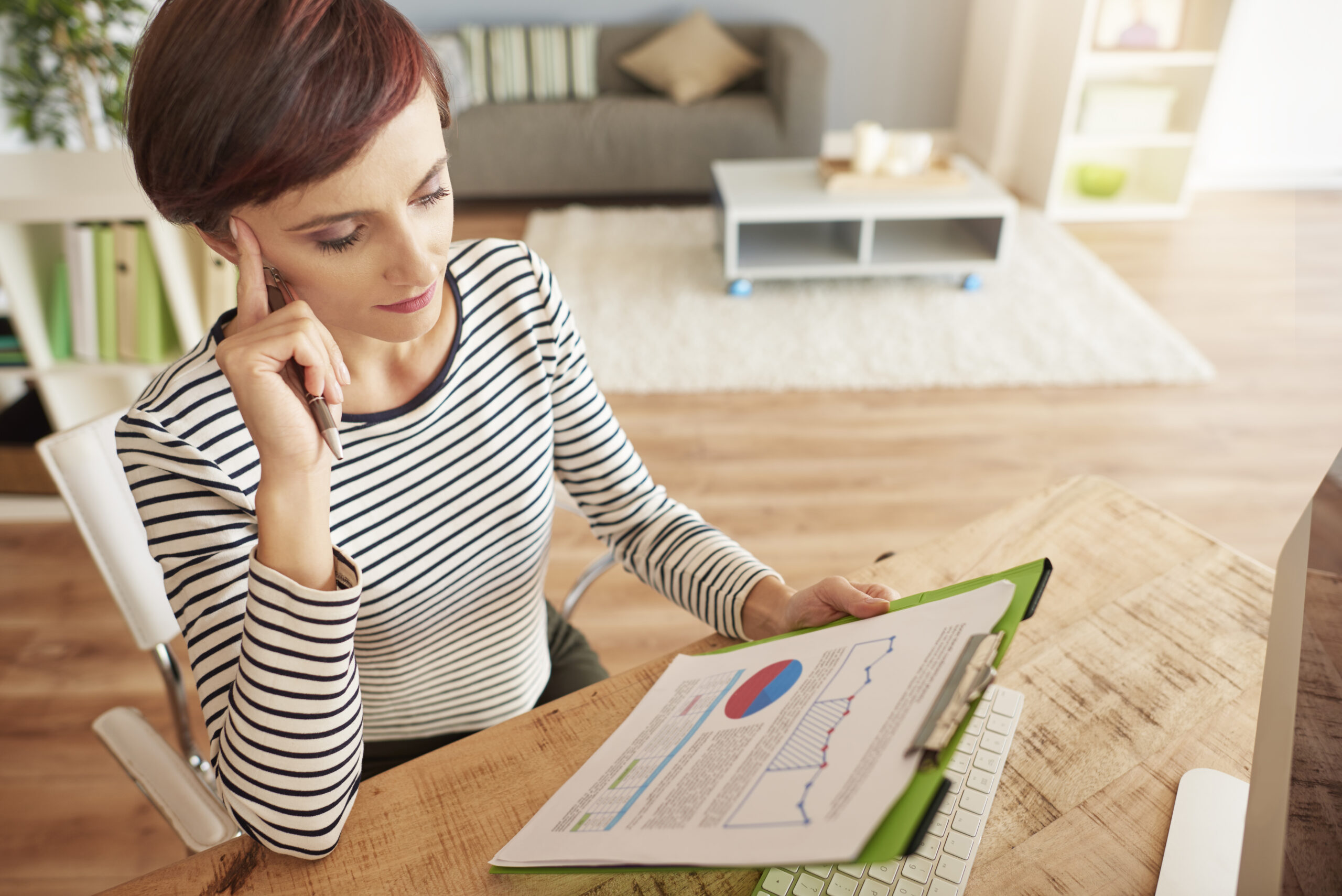 Woman in striped shirt sits at a desk, looking thoughtfully at documents with charts and graphs on a clipboard, in a home office setting.