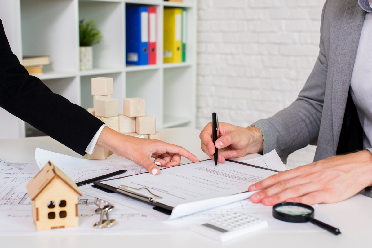 Two people review and sign documents at a desk with a small house model, wooden blocks, a magnifying glass, and office supplies.