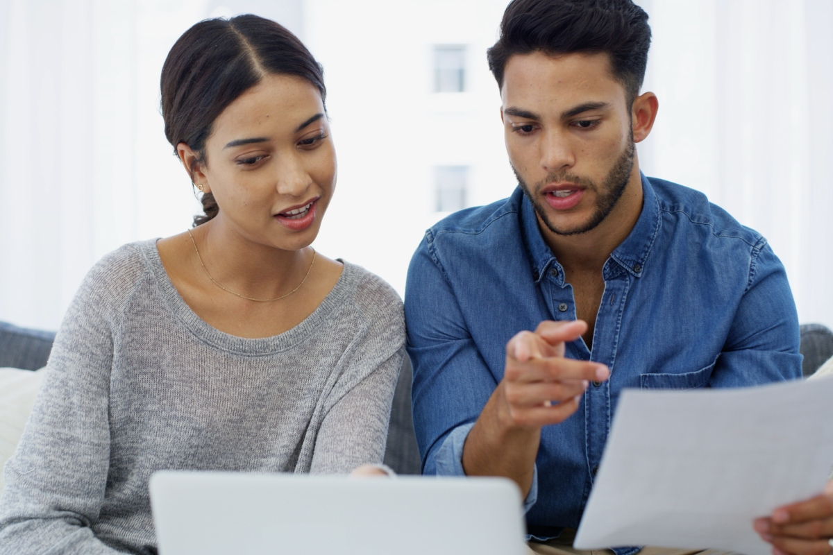 Two people sit together indoors, looking at a laptop and a document. One points at the paper while the other observes the screen.