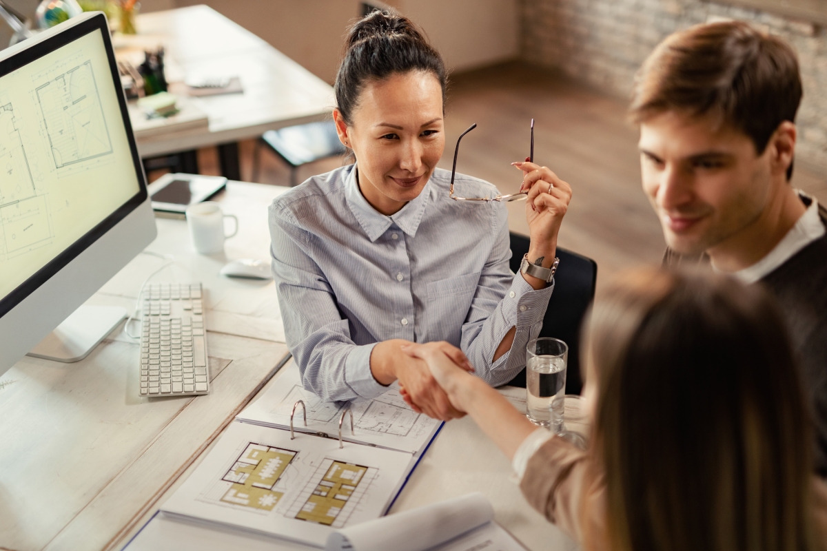 A woman in an office shakes hands with another woman across a desk with blueprints, while a man sits beside them.