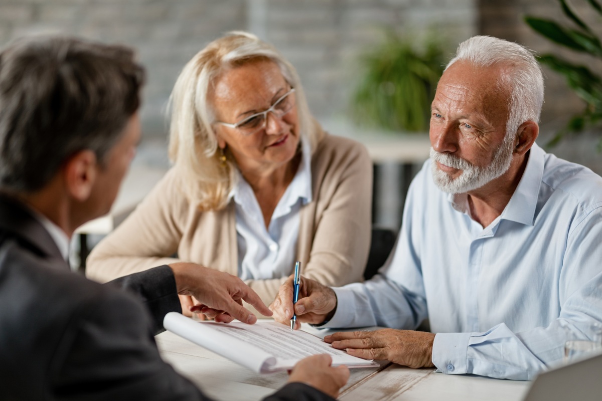 An elderly couple sits at a desk with a professional, reviewing and signing documents together in a bright, modern office setting.
